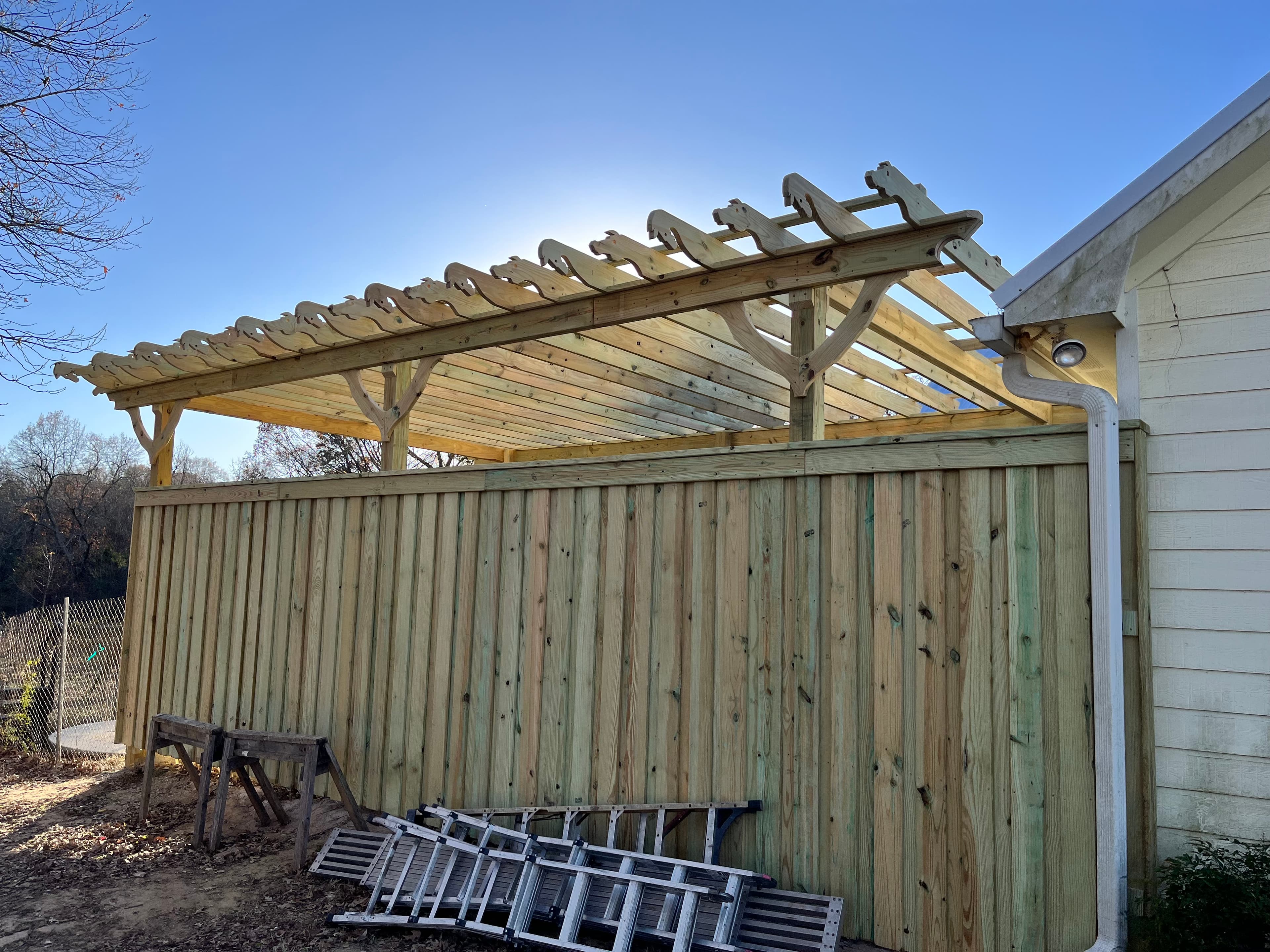 A wooden pergola with decorative rafter ends built atop a tall wooden privacy fence.